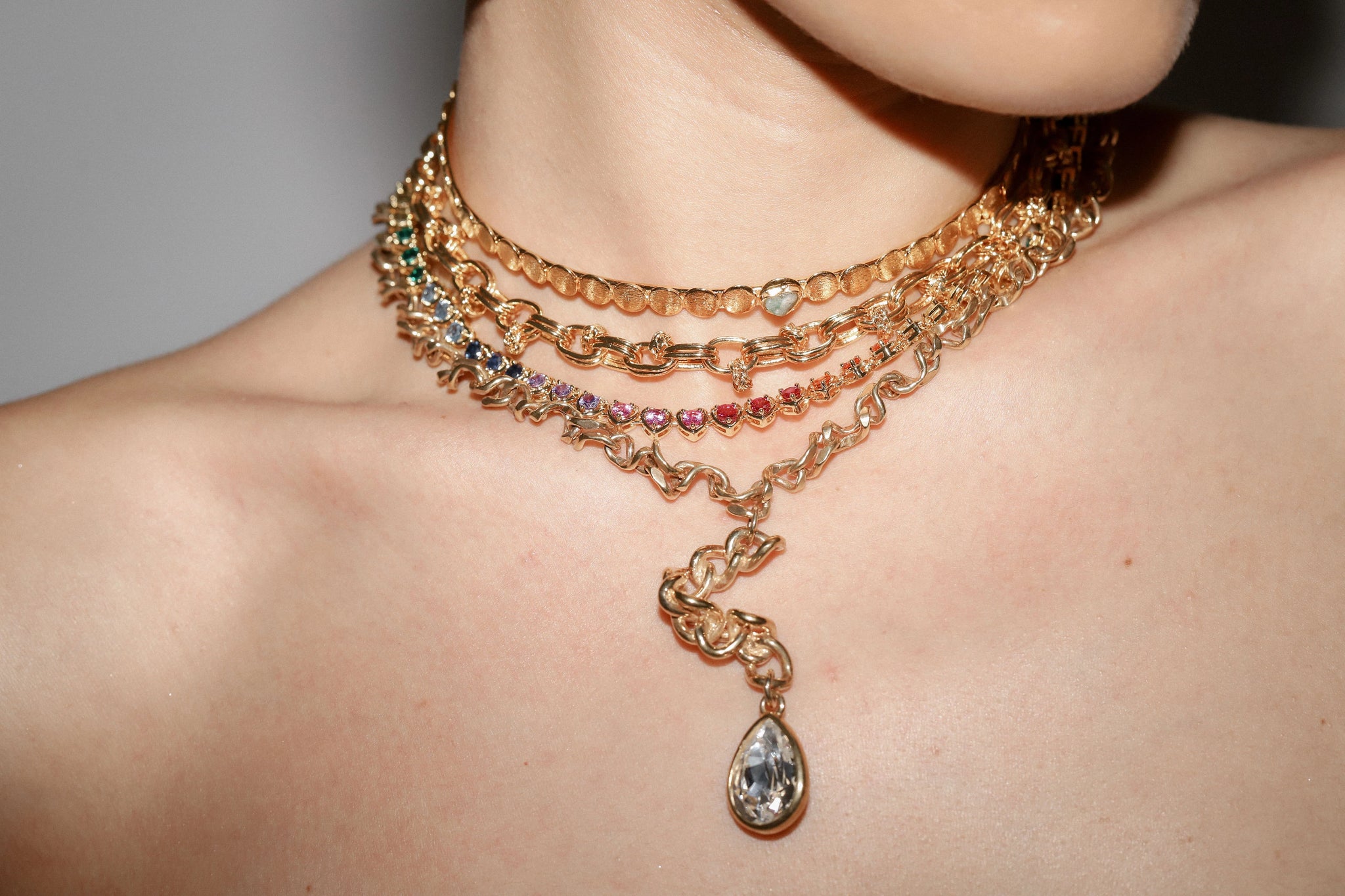 A close-up image of a woman’s neck adorned by unique, bejeweled gold necklaces.